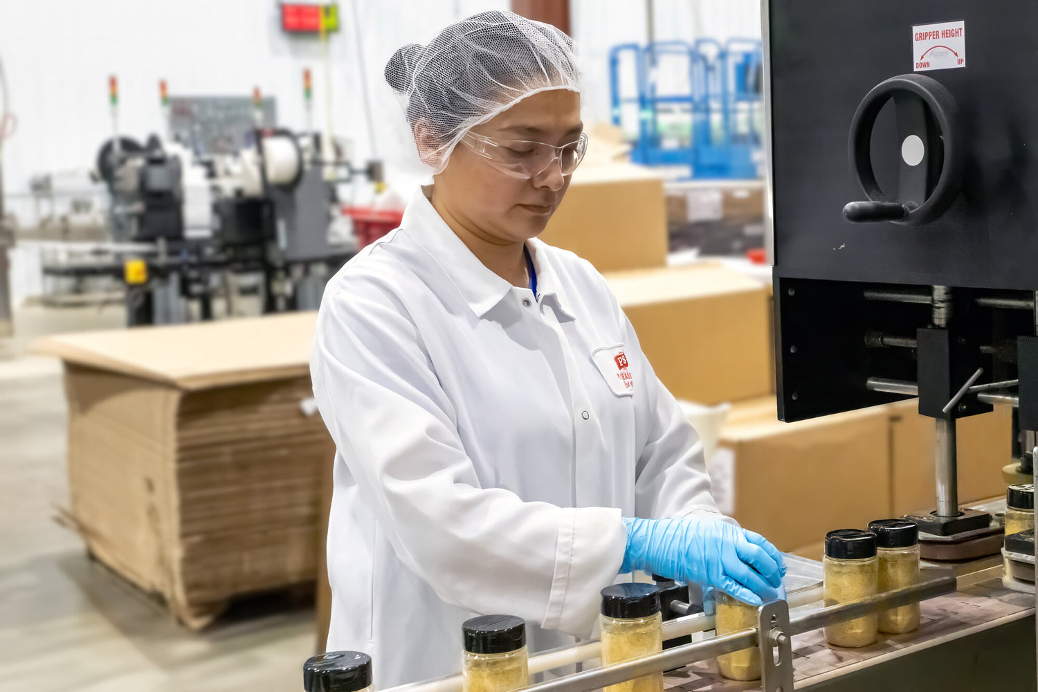 Person in a lab coat and hairnet working with jars in a factory setting