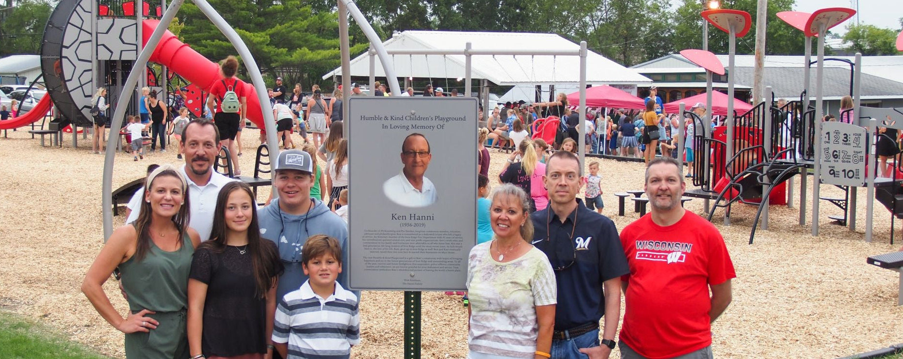 Group of people posing in front of a plaque with a photo of a man, likely at an outdoor event or park.